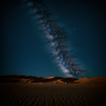 A vast alien desert landscape stretches under a mesmerizing blue nebula sky, showcasing an otherworldly and cosmic scene.