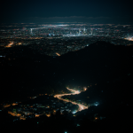 A stunning view of a city at night, seen from a mountain peak, showcasing twinkling city lights and a sprawling urban landscape under the starry sky.